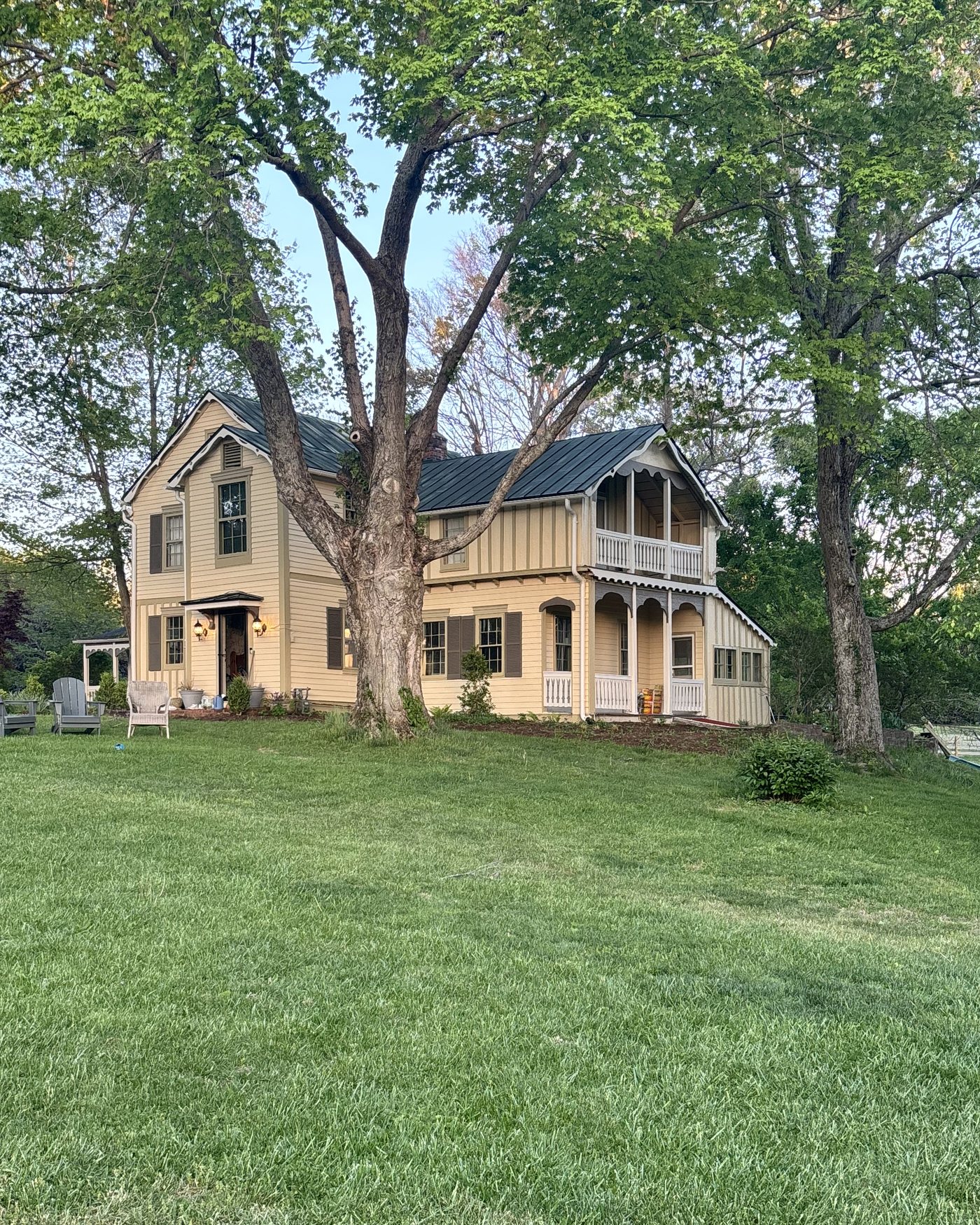 A yellow board-and-batten farmhouse with a gingerbread porch, under a large tree
